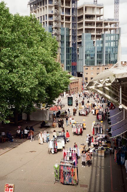 A busy outdoor market scene in an urban area with numerous vendor stalls set up on a paved pedestrian plaza, displaying various clothing and accessories such as dresses, jackets, and handbags. The market is adjacent to a large, leafy green tree on the left, providing shade, and is bordered by modern high-rise buildings with glass facades under construction in the background. Several shoppers and pedestrians are browsing and walking through the market, with some seated on benches along the left side. To the right, an overhanging structure with a metallic roof provides shelter for part of the market. The surroundings include a mix of construction activity and completed commercial developments, indicating a vibrant city center environment. This scene highlights alternative waste handling and private collection activities often associated with such outdoor markets, where vendors and shoppers might require rubbish removal services from companies like wasteclearanceelephantandcastle.co.uk to manage waste efficiently in busy urban settings.