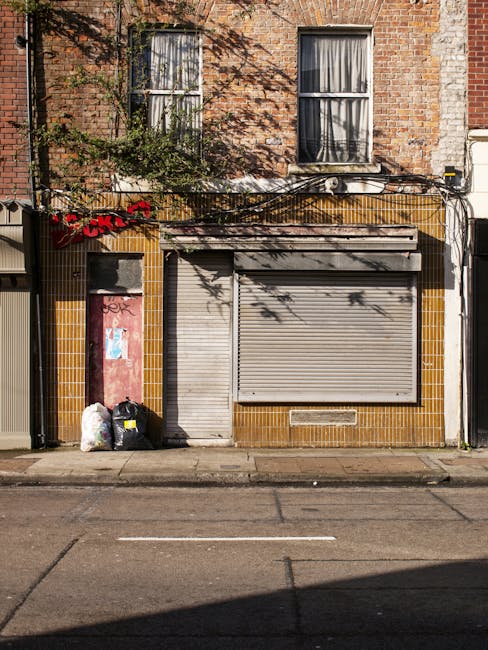 The image depicts the front of a small commercial or retail unit situated on a city street, with a closed, grey metal roller shutter covering the entrance and a matching smaller door to its left. The building exterior features a combination of brownish-yellow and beige tiles, with a weathered appearance. Above the storefront, the upper part of the building is constructed of red brick, with two rectangular sash windows framed in white, partially obscured by shadows and tree branches. To the left of the entrance, there are three black rubbish bags and two clear plastic bags filled with waste, placed on the sidewalk next to a pink-painted door that shows signs of graffiti. The sidewalk and street pavement are visible in the foreground with subtle cracks and markings, indicating regular urban wear. Overhead, a tangle of black electrical cables runs horizontally, partly casting shadows on the brick wall. This scene is typical of an independent waste collection or rubbish clearance service operating in a densely built urban environment, exemplifying private waste handling outside of municipal collection systems.