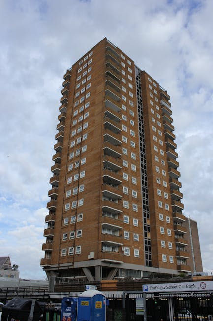 A tall, multi-storey residential building constructed from reddish-brown brick, featuring numerous small, rectangular windows with white frames arranged uniformly across each floor. The building has prominent balconies on the upper floors, with metal railings and concrete slabs, creating a repetitive pattern along the façade. The lower section includes a parking area enclosed by a black metal fence, with a visible sign indicating 'Trafalgar Street Car Park' and some off-road parking equipment such as blue portable toilets or storage units. The surrounding environment shows a partly cloudy sky with patches of blue, and the building stands upright in an urban setting, suggesting it is part of a densely populated area where independent waste collection or on-site clearance might be performed by specialists like wasteclearanceelephantandcastle.co.uk to manage rubbish removal from the property.