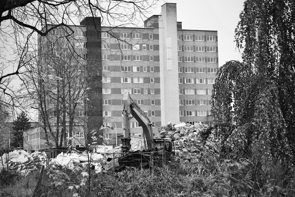 The image shows a construction or excavation site located in front of a multi-storey residential building with a grey brick exterior and numerous small, rectangular windows. In the foreground, there is an excavator with a long arm and bucket, positioned amidst a pile of broken concrete, debris, and scattered rocks. Surrounding the work area, there is overgrown vegetation, including tall grasses and leafy bushes. To the left, leafless trees with sprawling branches add a stark contrast to the scene, while on the right, a tree with dense foliage partially obscures some of the debris. The background features the apartment building, which has a flat roof and appears to be several stories high, with a uniform design. The overall environment appears to be a private site undergoing development or clearance work, consistent with independent waste handling or rubbish removal activities typical in urban areas seeking alternative disposal or clearance options, with the presence of machinery indicating ongoing bulk waste removal by a professional service like wasteclearanceelephantandcastle.co.uk.