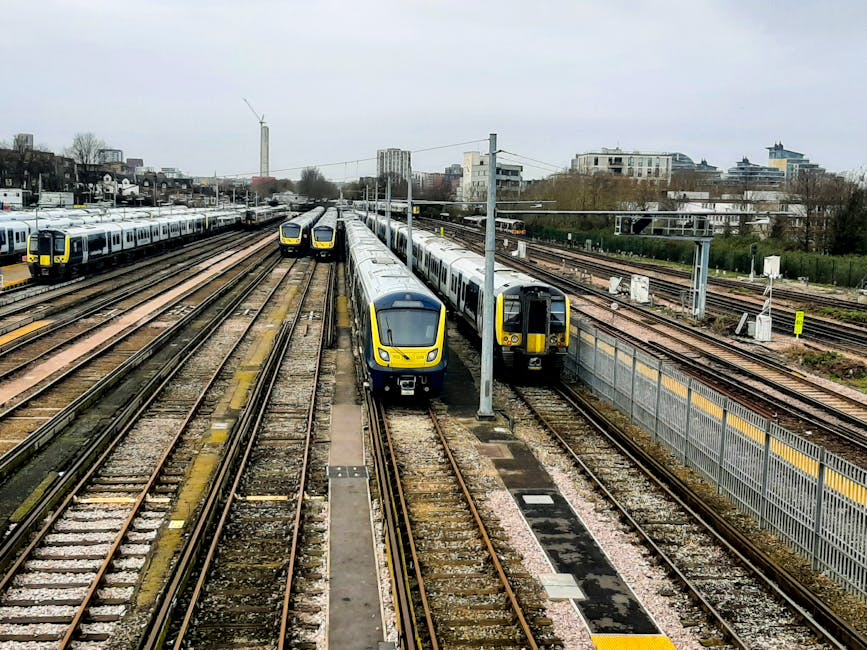 A railway yard featuring multiple train tracks with several modern passenger trains in yellow and gray livery parked on different lines. In the foreground, two trains are side by side—one facing forward and the other with its side visible—both with sleek, curved front designs and smooth metallic finishes. The tracks are constructed from steel rails on wooden sleepers, embedded in gravel, and appear weathered. Overhead, electrical wiring and supporting poles run parallel to the tracks, with some wires suspended above. In the background, there are additional train carriages, some stacked or lined up, along with a cityscape of mid-rise buildings, trees, and a cloudy sky. The environment appears to be an urban railway storage or maintenance area, with fences and safety barriers separating the tracks from adjacent spaces. Natural lighting is diffuse due to the overcast sky, highlighting the reflective surfaces of the trains and the textures of the gravel and infrastructure, subtly emphasizing the scene's functional aspect related to transport and railway operations, which aligns with the themes of alternative rail-based transportation and logistical management.