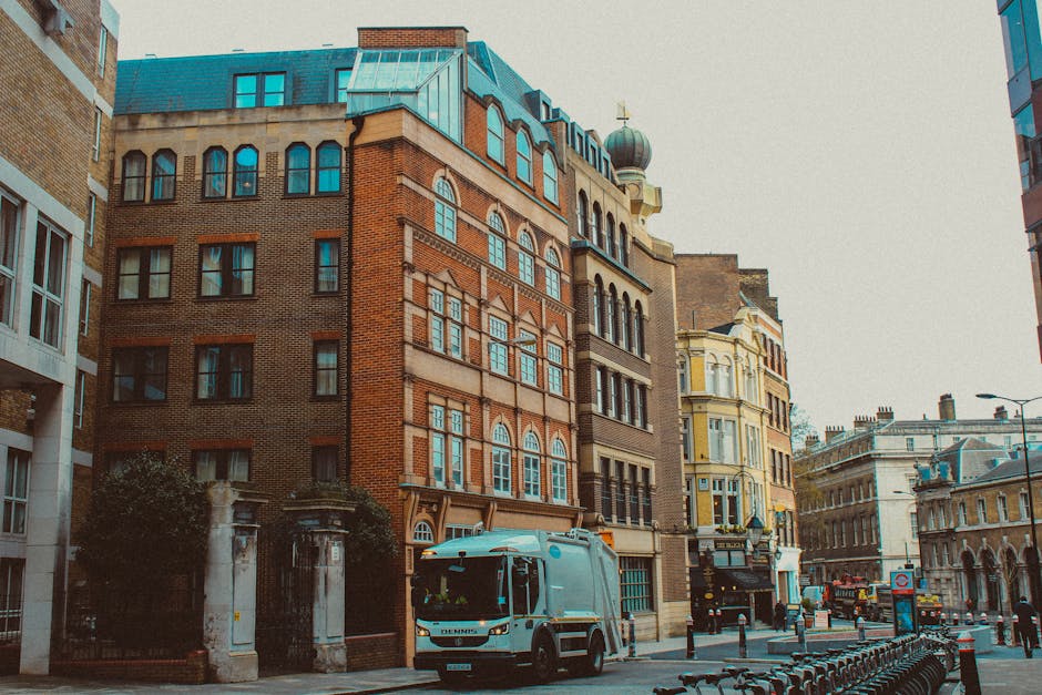 A city street scene featuring a row of multi-storey buildings with historical architectural styles, including a prominent red-brick structure with arched windows and a greenish copper roof. Adjacent buildings display various façade materials such as bricks, painted stucco, and decorative cornices. In the foreground, a light blue refuse collection vehicle with the company name (not visible) is parked on the pavement, part of a rubbish removal service. The street is lined with metal bike racks and bollards, with a few pedestrians and vehicles visible in the background. The environment appears to be an urban area with a mix of modern and older architecture, and the overall lighting suggests an overcast day, creating a neutral, professional atmosphere. This scene relates to the local alternative waste handling and on-site rubbish collection services provided by waste clearance specialists in a central London setting.