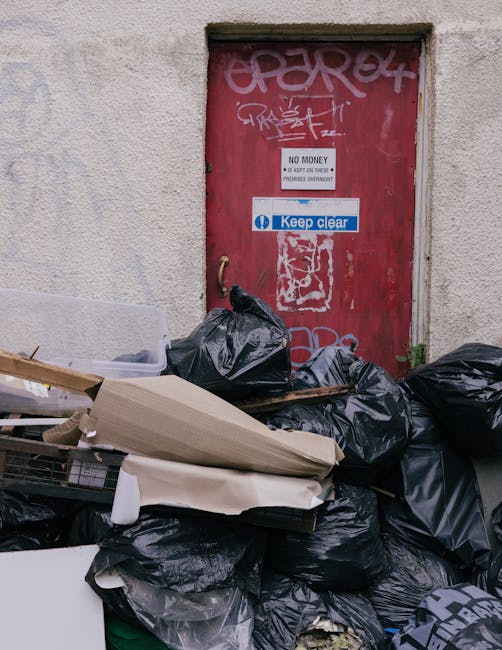 A large pile of black rubbish bags and discarded cardboard boxes stacked against a weathered red metal door set into a light-colored concrete wall. The bags appear to be filled with household waste or debris, with some partially torn open revealing contents. A few pieces of packaging and flattened cardboard are visible on top, leaning against the bags. The red door has graffiti tags and various signs attached; one sign indicates 'No Money' with additional smaller warning notices, and a blue and white sticker states 'Keep clear.' The environment suggests an alleyway or backstreet area, possibly used for informal waste disposal outside a residential or commercial building, consistent with private rubbish collection services. The scene is illuminated with natural daylight, highlighting the textures of the concrete wall, graffiti, and the crumpled plastic bags, emphasizing the need for effective waste clearance solutions such as those offered by waste management services like wasteclearanceelephantandcastle.co.uk.
